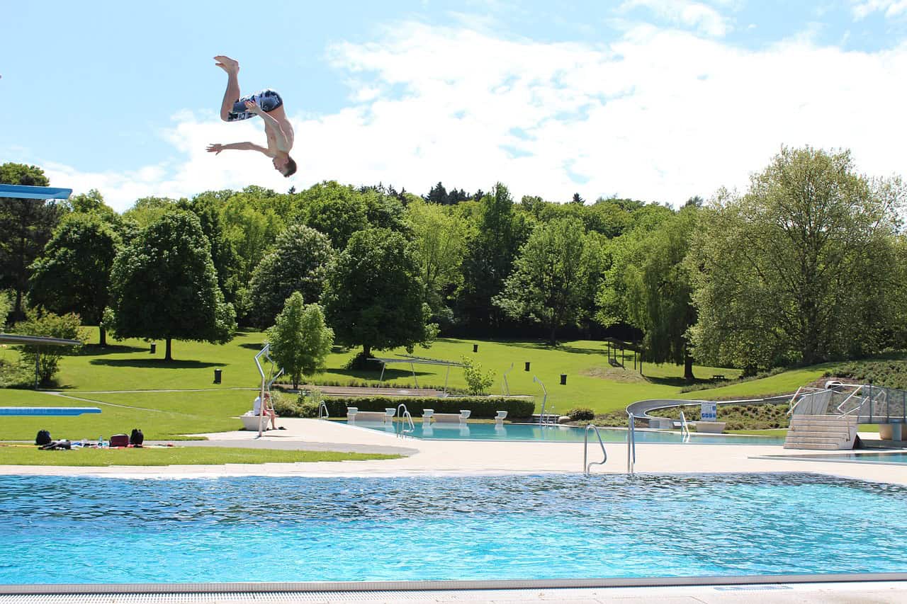 Vast swimming pool with a person diving into water, surrounded by lush green trees and sunny sky.